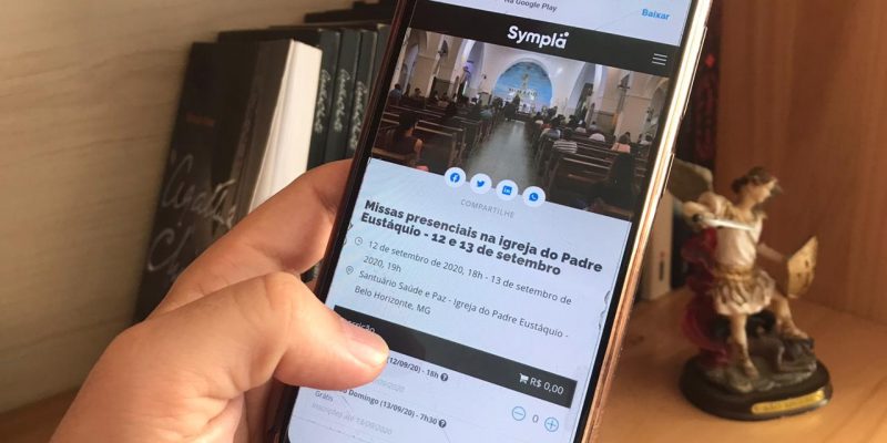 Mão com celular mostra a página do Sympla para retirada de ingressos para as missas presenciais.